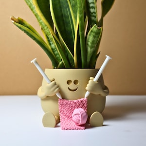 May include: A smiling planter shaped like a person holding knitting needles and a pink ball of yarn. The planter is holding a pink knitted piece of fabric.