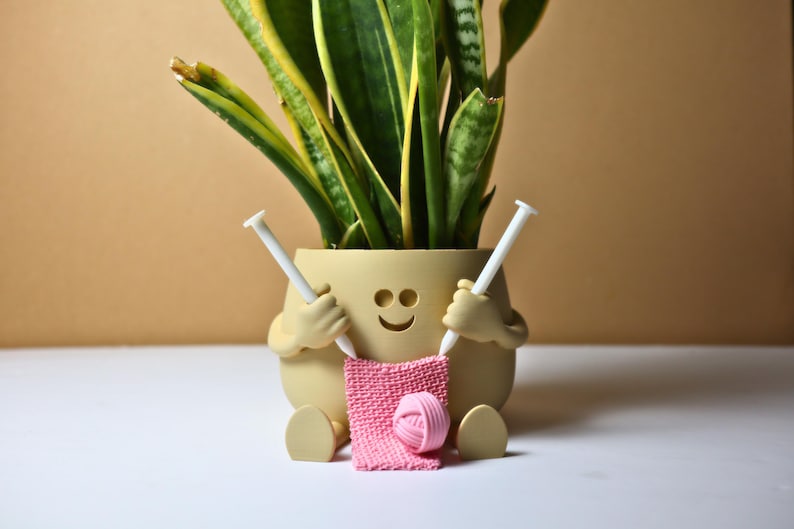 May include: A smiling planter shaped like a person holding knitting needles and a pink ball of yarn. The planter is holding a pink knitted piece of fabric.