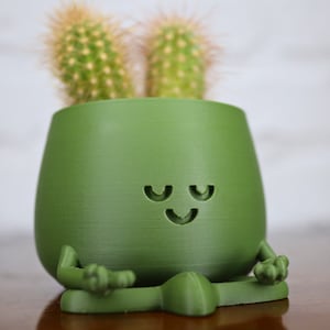 Può includere: Un vaso di ceramica verde a forma di figura che medita con un viso sorridente. Due piccole piante di cactus sono nel vaso.