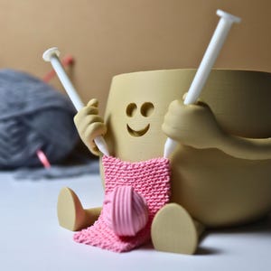 May include: A beige ceramic figurine of a person knitting with two white knitting needles. The figurine has a smiling face and is holding a pink knitted scarf. A ball of gray yarn is in the background.