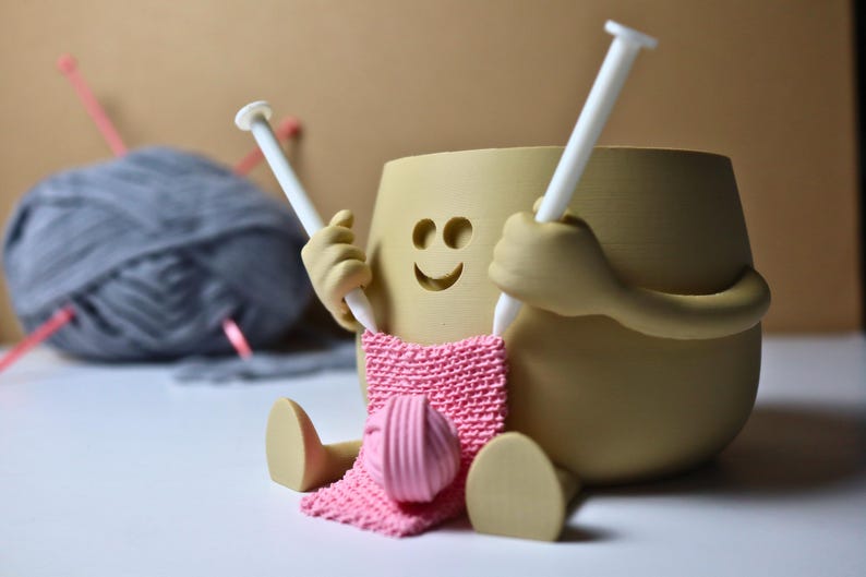 May include: A beige ceramic figurine of a person knitting with two white knitting needles. The figurine has a smiling face and is holding a pink knitted scarf. A ball of gray yarn is in the background.