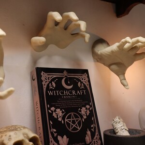 May include: A black book titled "Witchcraft: 4 Books in 1" with a pentagram and crescent moon design. The book is surrounded by three white ceramic hands, a lit candle, and a skull.