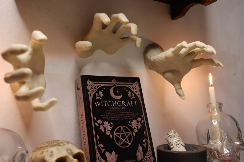 May include: A black book titled "Witchcraft: 4 Books in 1" with a pentagram and crescent moon design. The book is surrounded by three white ceramic hands, a lit candle, and a skull.