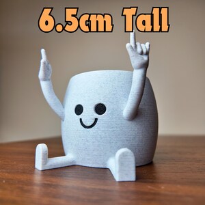 以下が含まれることがあります： 笑顔の人形の形をした、白い3Dプリントのプランター。腕を上げています。プランターの高さは6.5cmです。
