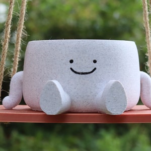 May include: A white ceramic planter shaped like a smiling character with two arms and legs, hanging from a rope swing. The planter is sitting on a red wooden platform.