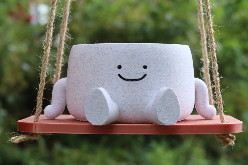 May include: A white ceramic planter shaped like a smiling character with two arms and legs, hanging from a rope swing. The planter is sitting on a red wooden platform.