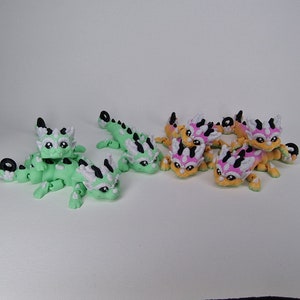 Puede incluir: Un grupo de peque&ntilde;as figuritas de drag&oacute;n articuladas impresas en 3D. Los dragones est&aacute;n en dos esquemas de color: verde con detalles blancos y naranja con detalles rosas y blancos.