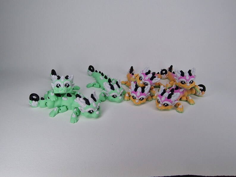 Puede incluir: Un grupo de peque&ntilde;as figuritas de drag&oacute;n articuladas impresas en 3D. Los dragones est&aacute;n en dos esquemas de color: verde con detalles blancos y naranja con detalles rosas y blancos.