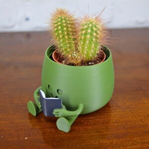 Puede incluir: Un macetero de cer&aacute;mica verde con forma de persona sentada con las piernas cruzadas y sosteniendo un libro. El macetero tiene una planta de cactus creciendo dentro.