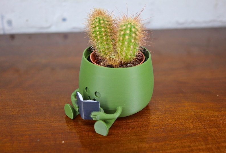 Puede incluir: Un macetero de cer&aacute;mica verde con forma de persona sentada con las piernas cruzadas y sosteniendo un libro. El macetero tiene una planta de cactus creciendo dentro.