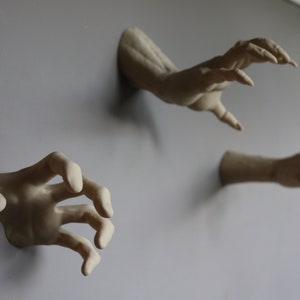 May include: Three plaster hands reaching out from a wall, each with a different pose. The hands are painted in a light beige color.