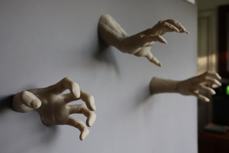 May include: Three plaster hands reaching out from a wall, each with a different pose. The hands are painted in a light beige color.