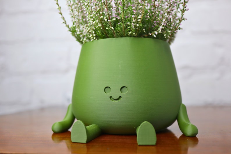 Puede incluir: Una maceta de cer&aacute;mica verde con una cara sonriente y peque&ntilde;as patas. La maceta est&aacute; llena de una peque&ntilde;a planta con flores blancas.