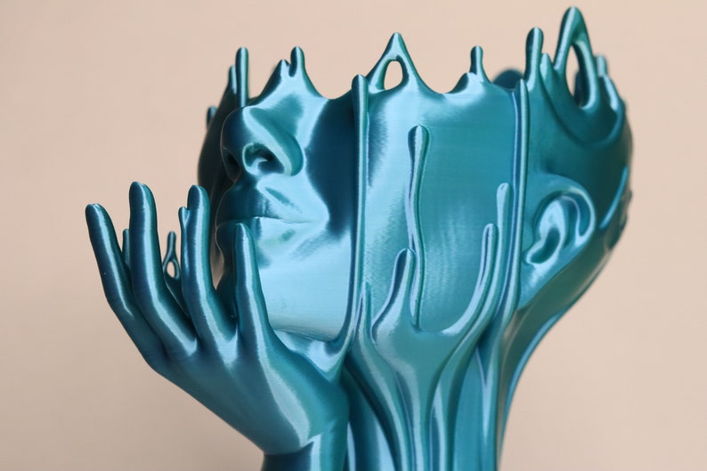 May include: A teal blue abstract sculpture of a face with hands reaching up. The sculpture is made of a smooth, flowing material that resembles liquid.