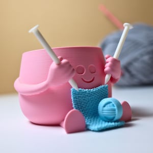 May include: A pink ceramic figurine of a person knitting with two white knitting needles and a blue ball of yarn. The figurine has a smiling face and is holding the needles in its hands.