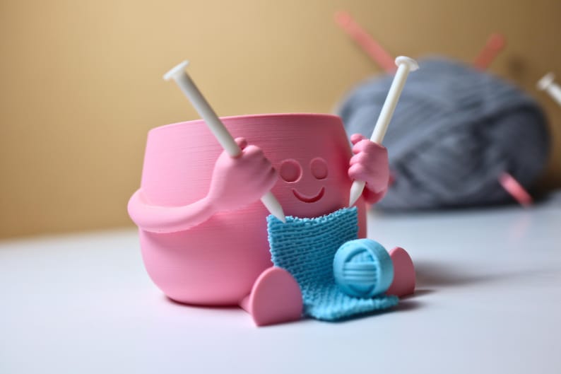 May include: A pink ceramic figurine of a person knitting with two white knitting needles and a blue ball of yarn. The figurine has a smiling face and is holding the needles in its hands.