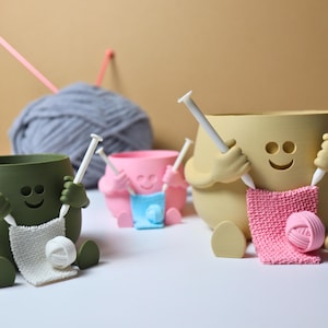 May include: Three ceramic yarn bowls shaped like smiling faces with arms and legs. Each bowl has a pair of knitting needles sticking out of its hands and a ball of yarn in its lap. The bowls are green, yellow, and pink.