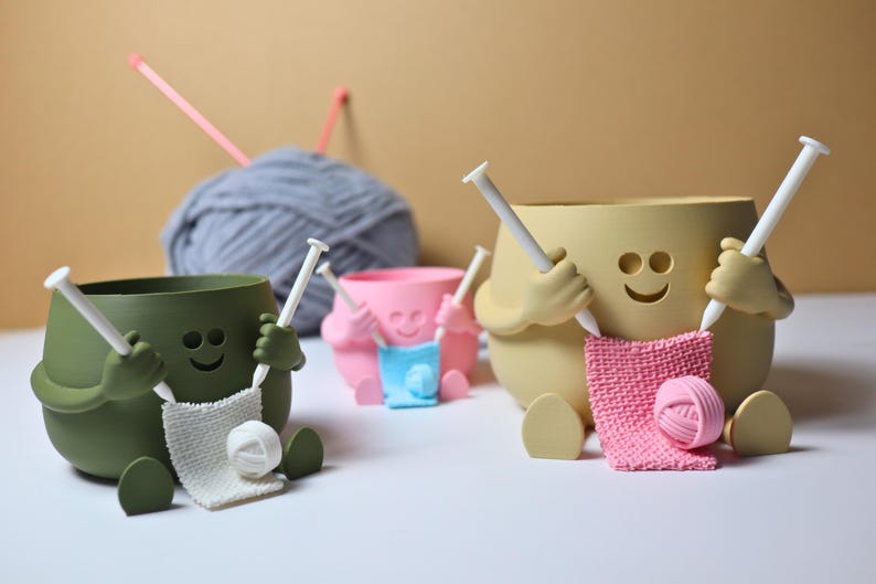 May include: Three ceramic yarn bowls shaped like smiling faces with arms and legs. Each bowl has a pair of knitting needles sticking out of its hands and a ball of yarn in its lap. The bowls are green, yellow, and pink.