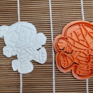 May include: An orange plastic cookie cutter and a white cookie in the shape of a cartoon character. The cutter is in the shape of a bear with geometric features. The cookie is a solid white color and has the same bear shape.