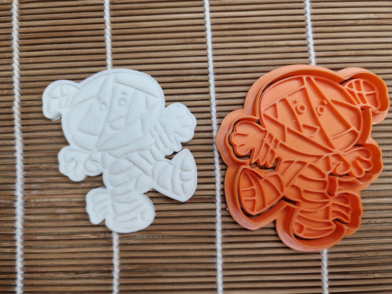May include: An orange plastic cookie cutter and a white cookie in the shape of a cartoon character. The cutter is in the shape of a bear with geometric features. The cookie is a solid white color and has the same bear shape.
