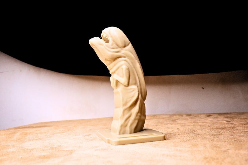 May include: A beige plastic figurine of a creature with a large mouth and a hooded head. The creature is standing on a small beige plastic base.