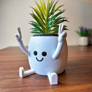 May include: A whimsical, light gray planter with a smiling face, arms raised in a peace sign, and legs. The planter holds a green and yellow artificial plant. The planter is sitting on a brown wooden surface.