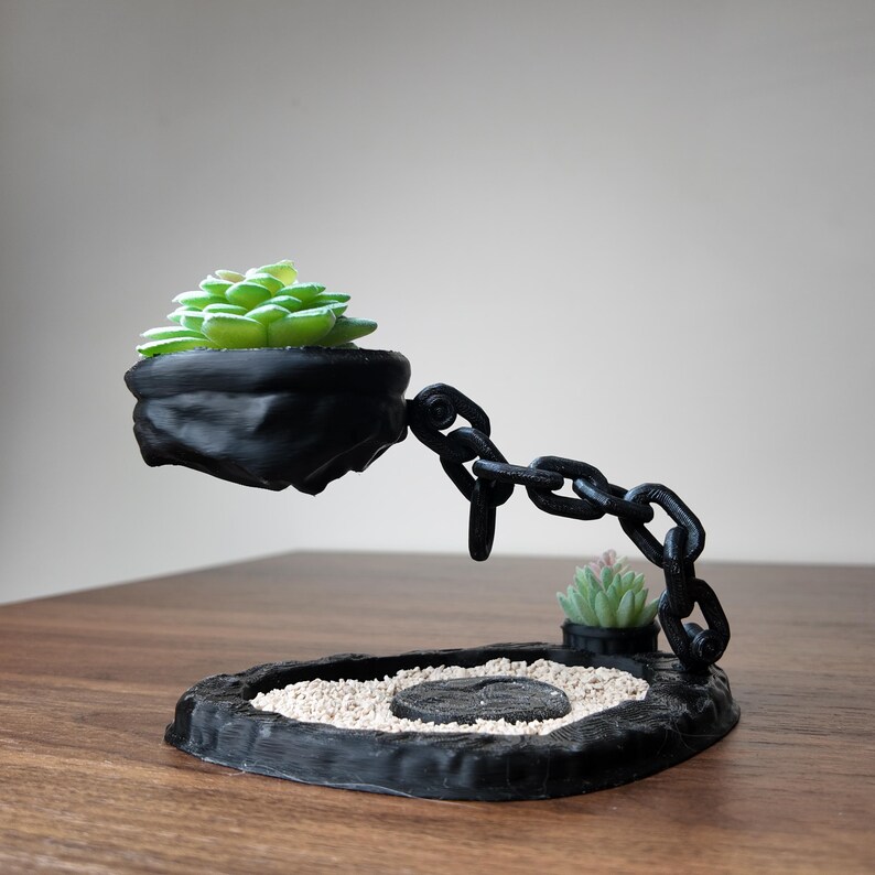 May include: A black, 3D-printed succulent planter with a chain design. The planter holds a green succulent and is suspended above a base with a small succulent and white sand. The base is black and has a zen garden aesthetic.