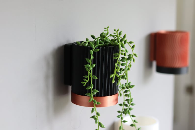 May include: A black and copper wall planter with a green succulent plant. The planter has a ribbed design and a copper band around the top.