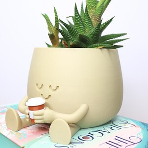 May include: A beige ceramic planter shaped like a person sitting with legs crossed, holding a small coffee cup. The planter has a smiling face and closed eyes. A green succulent plant is growing inside the planter.