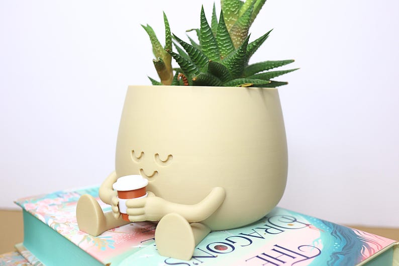 May include: A beige ceramic planter shaped like a person sitting with legs crossed, holding a small coffee cup. The planter has a smiling face and closed eyes. A green succulent plant is growing inside the planter.