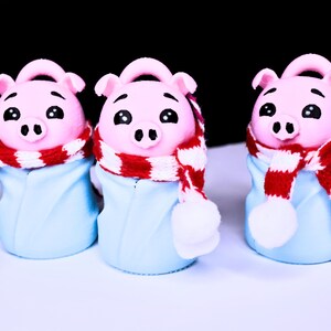 May include: Three pink pig figurines wearing blue and white winter outfits with red and white scarves.