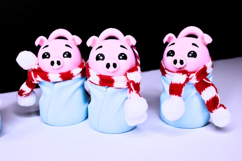 May include: Three pink pig figurines wearing blue and white winter outfits with red and white scarves.
