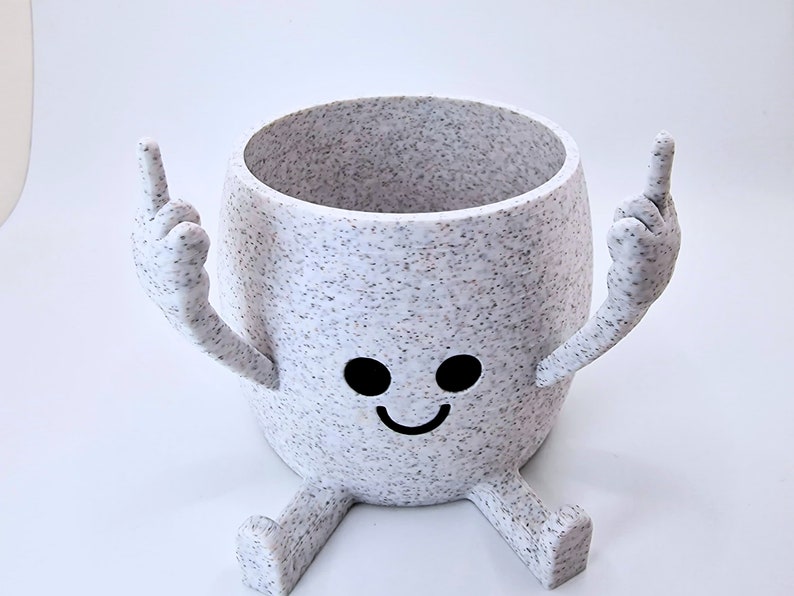 May include: White ceramic planter with a speckled finish, shaped like a person with two arms raised in the air and two legs. The planter has a smiling face with two black eyes.
