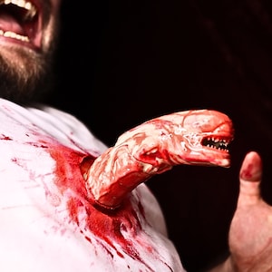 May include: A close-up of a person with a bloody white shirt, a creature bursting out of their chest, and a hand reaching out in horror.