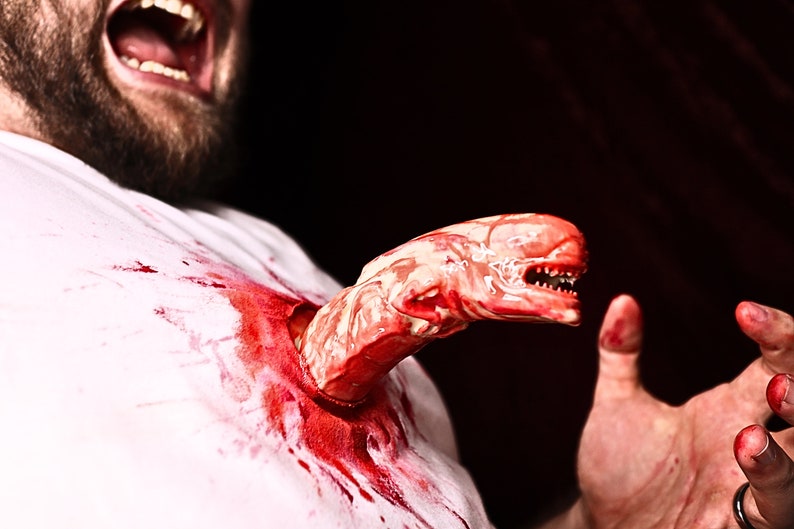 May include: A close-up of a person with a bloody white shirt, a creature bursting out of their chest, and a hand reaching out in horror.