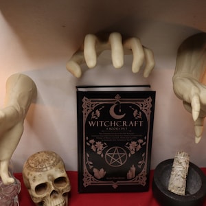 May include: A black book titled "Witchcraft: A Book in 1" with a pentagram and crescent moon design on the cover. The book is surrounded by white ceramic hands, a skull, a mortar and pestle, and a glass bottle.