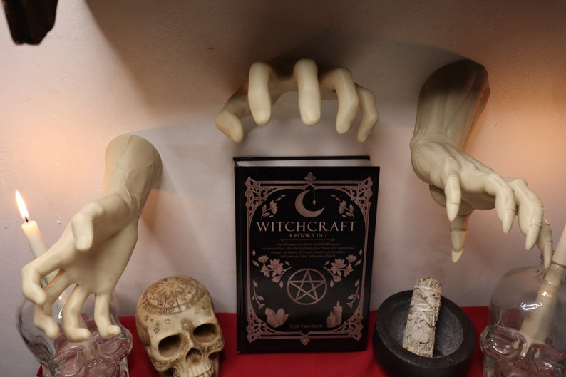 May include: A black book titled "Witchcraft: A Book in 1" with a pentagram and crescent moon design on the cover. The book is surrounded by white ceramic hands, a skull, a mortar and pestle, and a glass bottle.