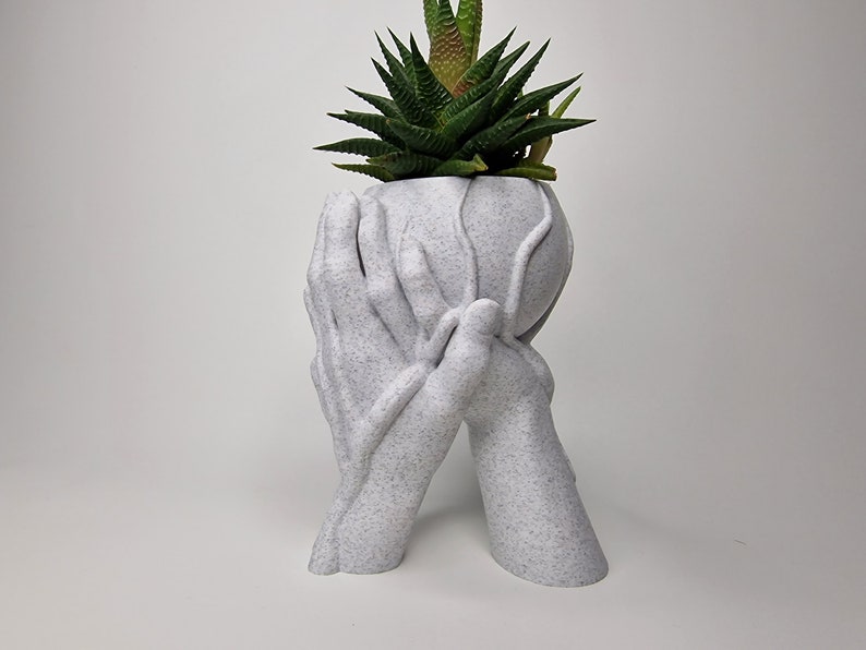 Puede incluir: Maceta de cer&aacute;mica blanca con forma de dos manos sosteniendo una esfera, con una planta suculenta verde creciendo en su interior.