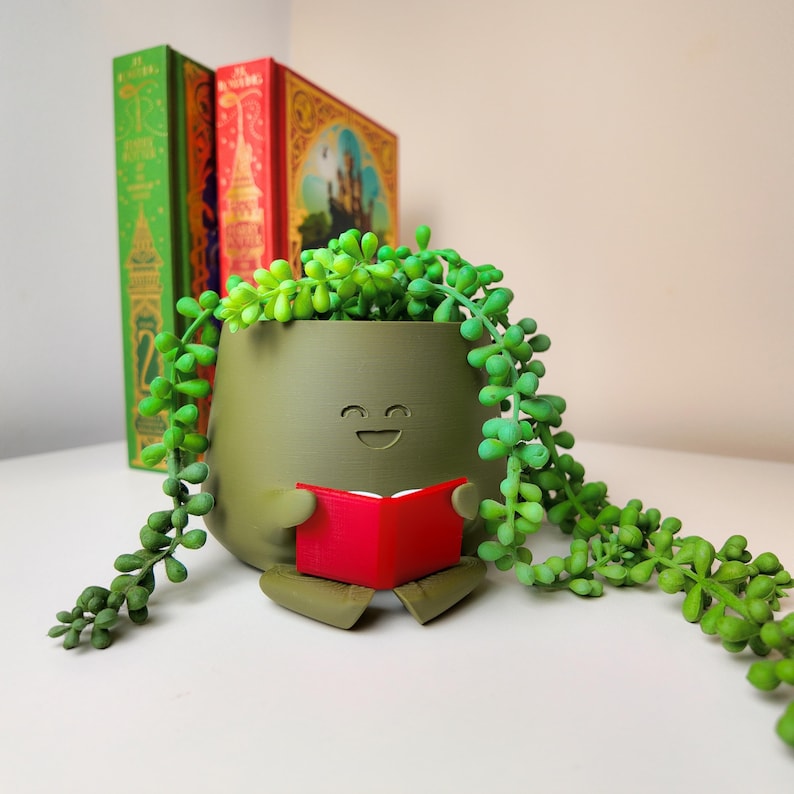 Puede incluir: Un macetero de cer&aacute;mica verde con forma de persona sentada con un libro rojo en sus manos. El macetero tiene una cara sonriente y est&aacute; lleno de una planta suculenta verde.