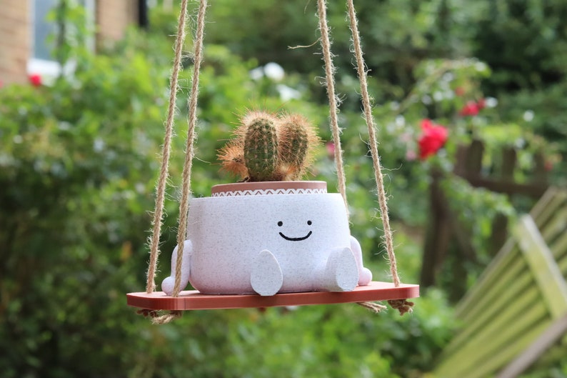 May include: A white ceramic planter with a smiling face and legs, hanging from a rope swing. The planter contains a small cactus plant.