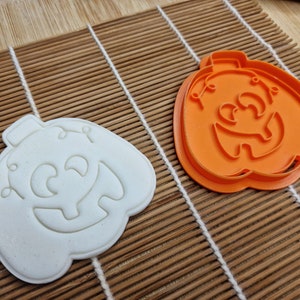May include: An orange plastic pumpkin-shaped cookie cutter and a white cookie in the same shape. The cutter has a jack-o'-lantern face design. The items are on a bamboo mat, perfect for Halloween baking.