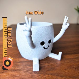 Puede incluir: Maceta de cer&aacute;mica gris claro, de 8 cm de alto y 8 cm de ancho, con forma de personaje de dibujos animados. Tiene una cara sonriente, brazos levantados en se&ntilde;al de paz y piernas. Ideal para plantas peque&ntilde;as.
