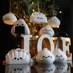 May include: A collection of white ceramic ornaments shaped like dumplings, onigiri, and bao buns. Each ornament has a cute cartoon face and is decorated with pink and red details. The ornaments are hanging from a branch and are arranged in front of a wooden sign that says "LOVE".