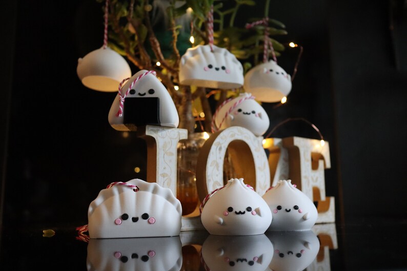 May include: A collection of white ceramic ornaments shaped like dumplings, onigiri, and bao buns. Each ornament has a cute cartoon face and is decorated with pink and red details. The ornaments are hanging from a branch and are arranged in front of a wooden sign that says "LOVE".