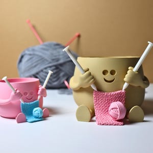 May include: Two ceramic yarn bowls shaped like smiling faces holding knitting needles and yarn. The bowl on the left is pink and the bowl on the right is beige.