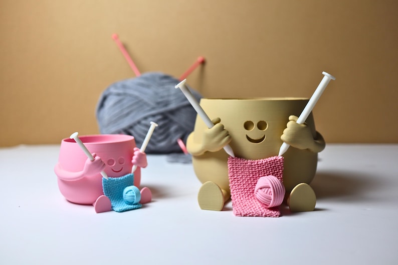 May include: Two ceramic yarn bowls shaped like smiling faces holding knitting needles and yarn. The bowl on the left is pink and the bowl on the right is beige.
