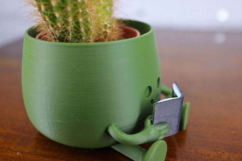 Puede incluir: Una maceta verde con forma de persona leyendo un libro. La maceta tiene agujeros de drenaje y est&aacute; hecha de un material texturizado. El cactus dentro de la maceta es verde y tiene muchas espinas.