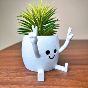 Puede incluir: Una maceta de cer&aacute;mica blanca moteada con una cara sonriente y brazos haciendo el signo de la paz. Contiene una suculenta artificial verde y amarilla. La maceta est&aacute; sobre una superficie de madera.