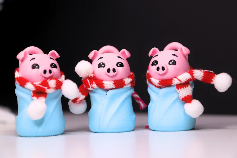 May include: Three pink pig figurines wearing blue and white winter outfits with red and white striped scarves. The pigs have black eyes and snouts.