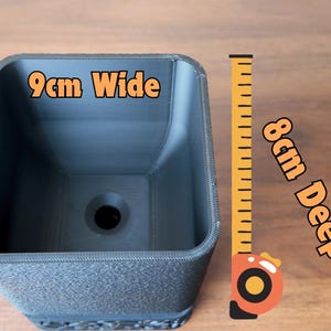 May include: A dark gray square planter, 9cm wide and 8cm deep, with a textured exterior. The planter has a drainage hole at the bottom. A yellow measuring tape is next to the planter.